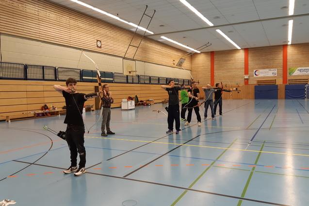 Sechs Bogensch&uuml;tzen spannen ihre B&ouml;gen in einer Sporthalle mit blauen Fu&szlig;bodenmarkierungen.