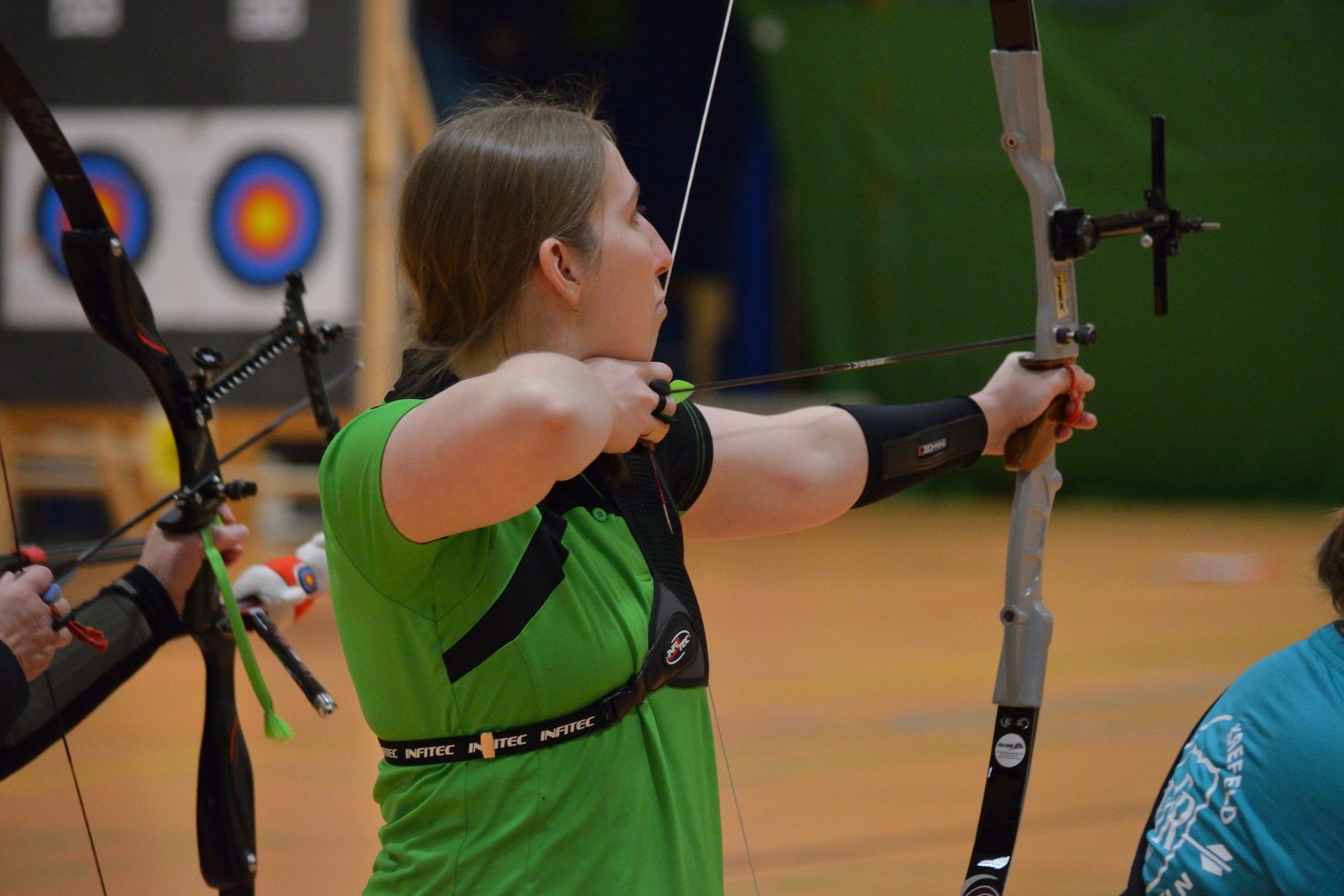 Eine Sportlerin in gr&uuml;ner Kleidung zieht einen Bogen in einer Halle, w&auml;hrend ein Ziel im Hintergrund sichtbar ist.