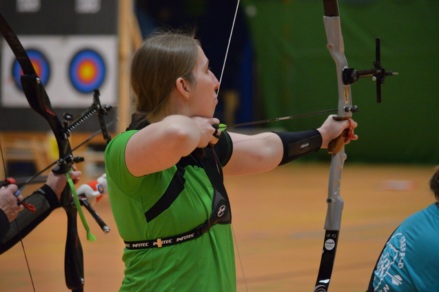 Eine Sportlerin in gr&uuml;ner Kleidung zieht einen Bogen in einer Halle, w&auml;hrend ein Ziel im Hintergrund sichtbar ist.