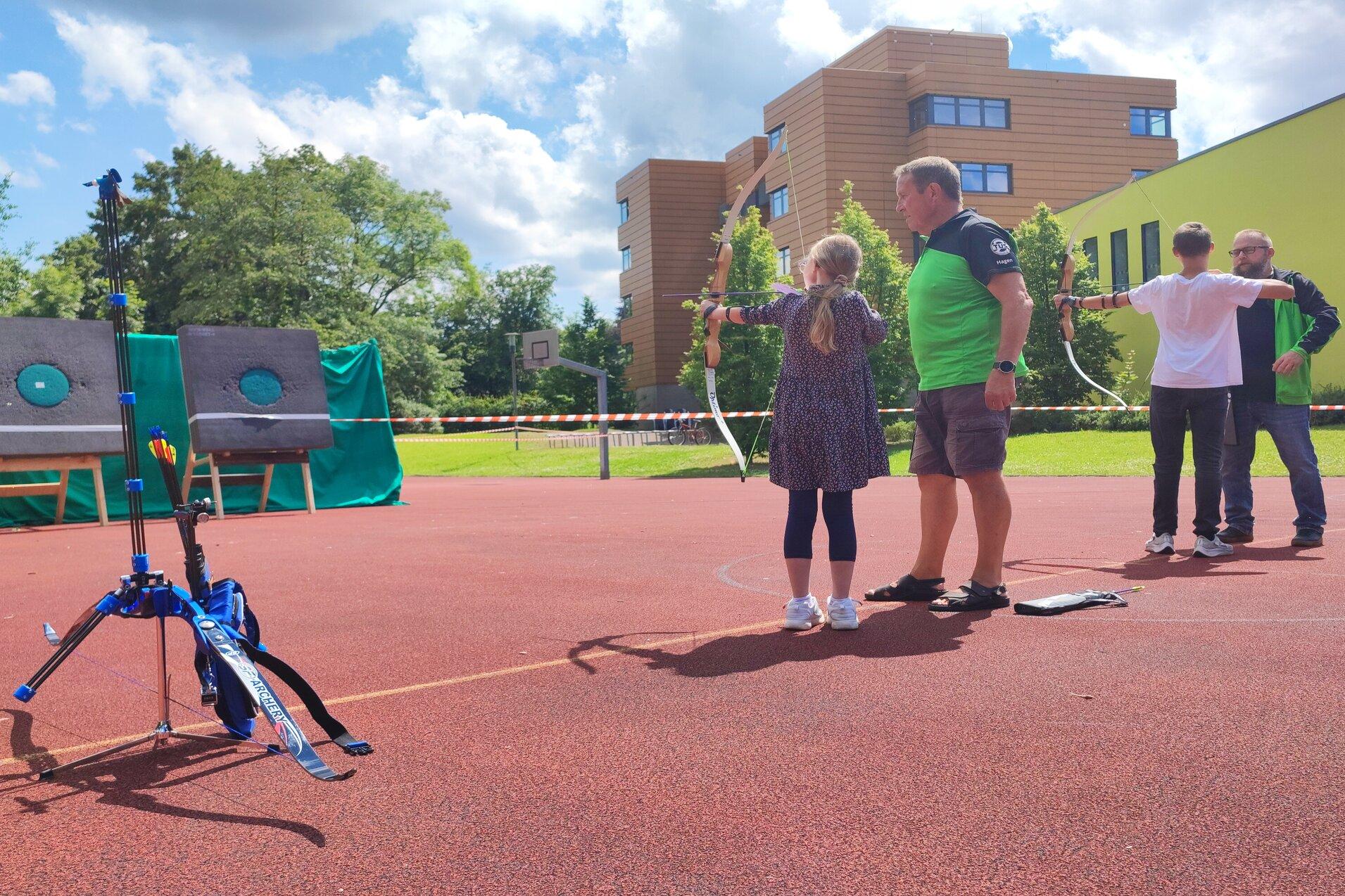 Eine Gruppe von Personen steht auf einem Sportplatz und &uuml;bt Bogenschie&szlig;en. Ziele sind in der Entfernung sichtbar.