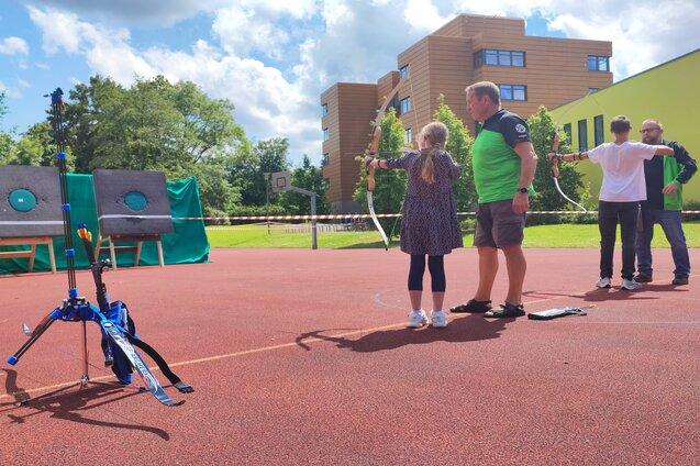 Eine Gruppe von Personen steht auf einem Sportplatz und &uuml;bt Bogenschie&szlig;en. Ziele sind in der Entfernung sichtbar.
