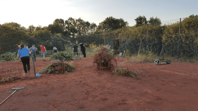 Menschen arbeiten gemeinsam beim Entfernen von Unkraut auf einem vernachlässigten Tennisplatz mit rotem Sandboden.