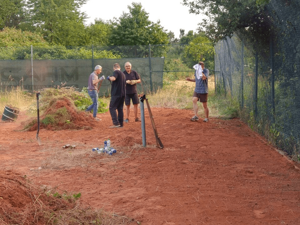 Eine Gruppe von vier Männern arbeitet im Freien und räumt eine sandige Fläche mit Werkzeugen und Pflanzenresten auf.