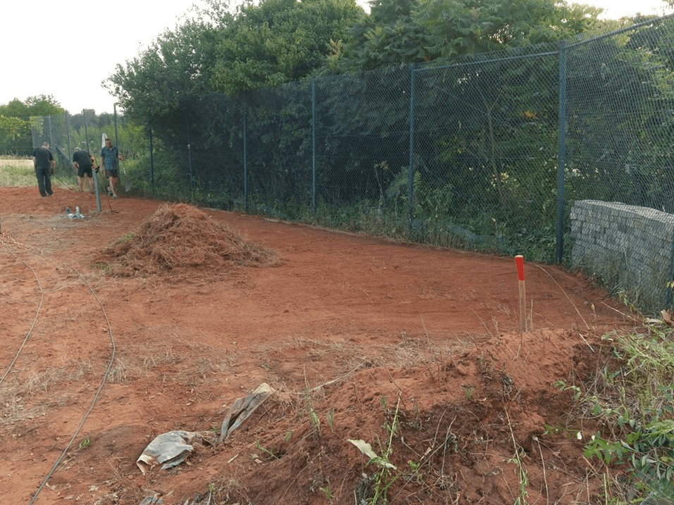 Bauarbeiten an einem Platz aus roter Erde, mit einem Erdhaufen und mehreren Personen in einem umzäunten Bereich.