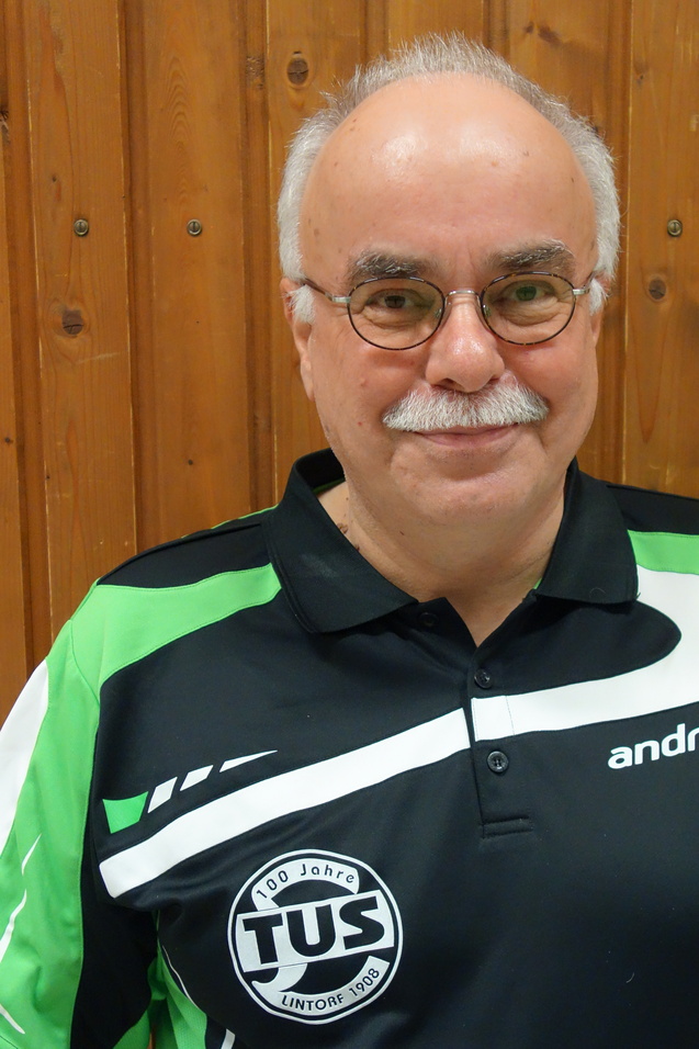 Ein &auml;lterer Mann mit Brille und Schnurrbart l&auml;chelt vor einer Holzwand, tr&auml;gt ein gr&uuml;nes Sportshirt mit TUS-Logo.