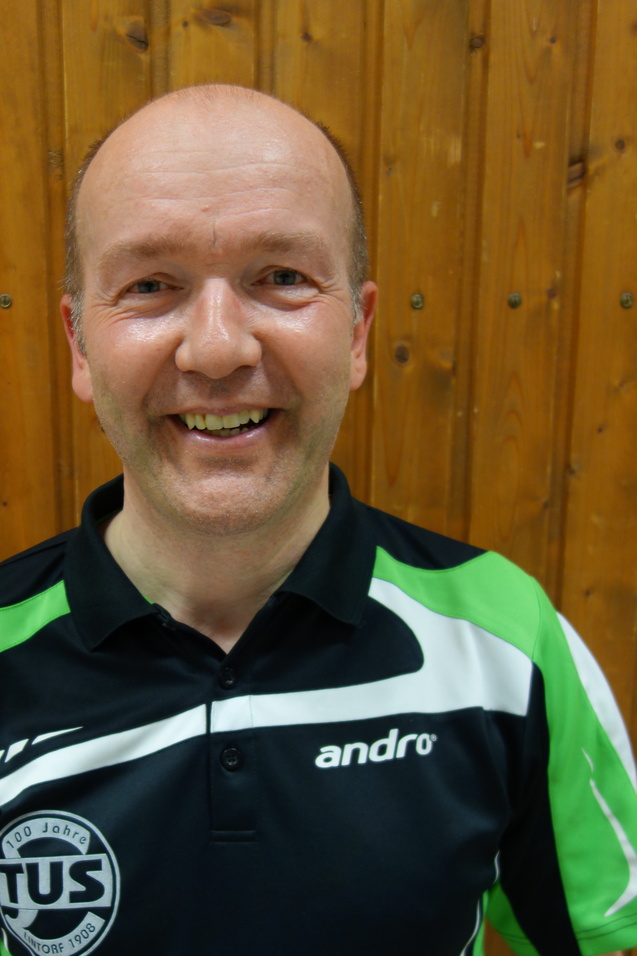 L&auml;chelnder Mann in einem schwarzen und gr&uuml;nen Polo-Hemd vor einer Holzvert&auml;felung, eventuell Tischtennistrainer.