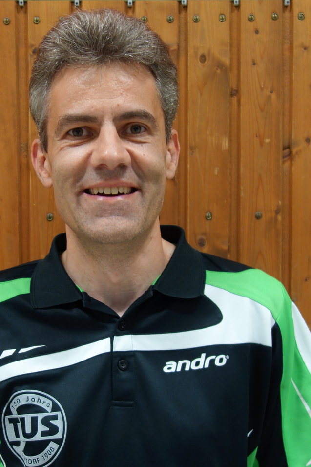 Portr&auml;t eines l&auml;chelnden Mannes in einem schwarzen Poloshirt mit gr&uuml;nen Akzenten vor einer Holzwand.