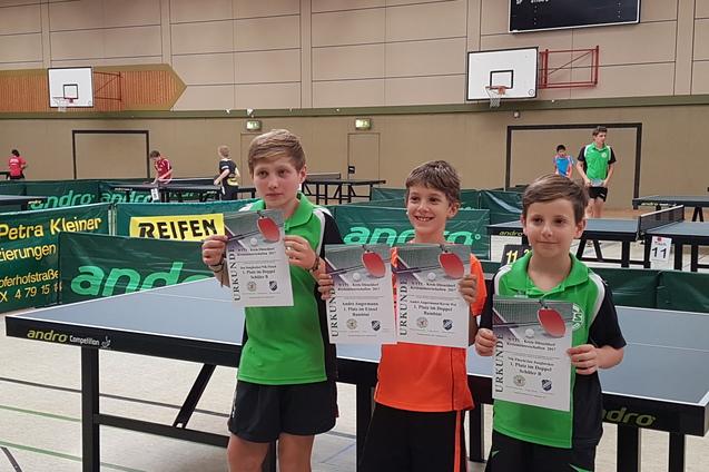 Drei Jungen halten Urkunden und Tischtennisschl&auml;ger in einer Sporthalle mit Tischtennisplatten im Hintergrund.