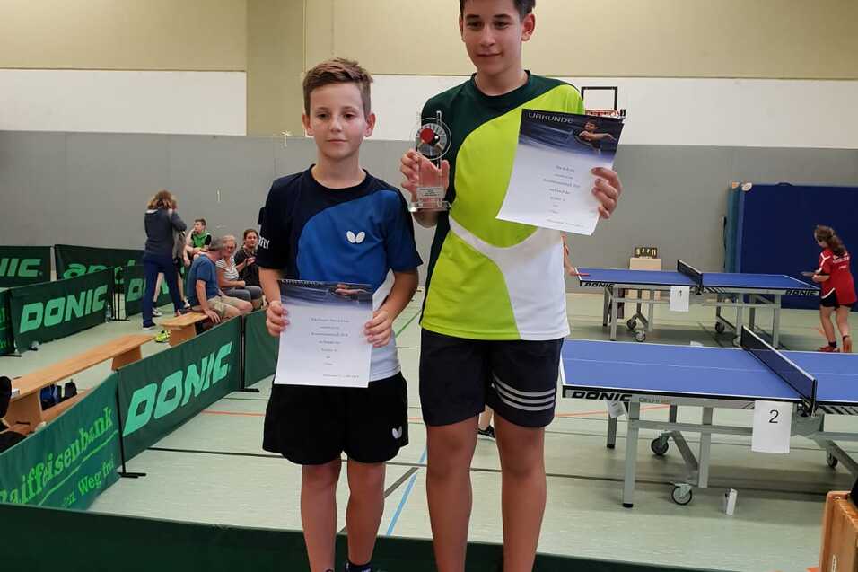 Zwei Kinder stehen auf dem Siegerpodest, halten Urkunden und einen Pokal, umgeben von Tischtennisplatten.