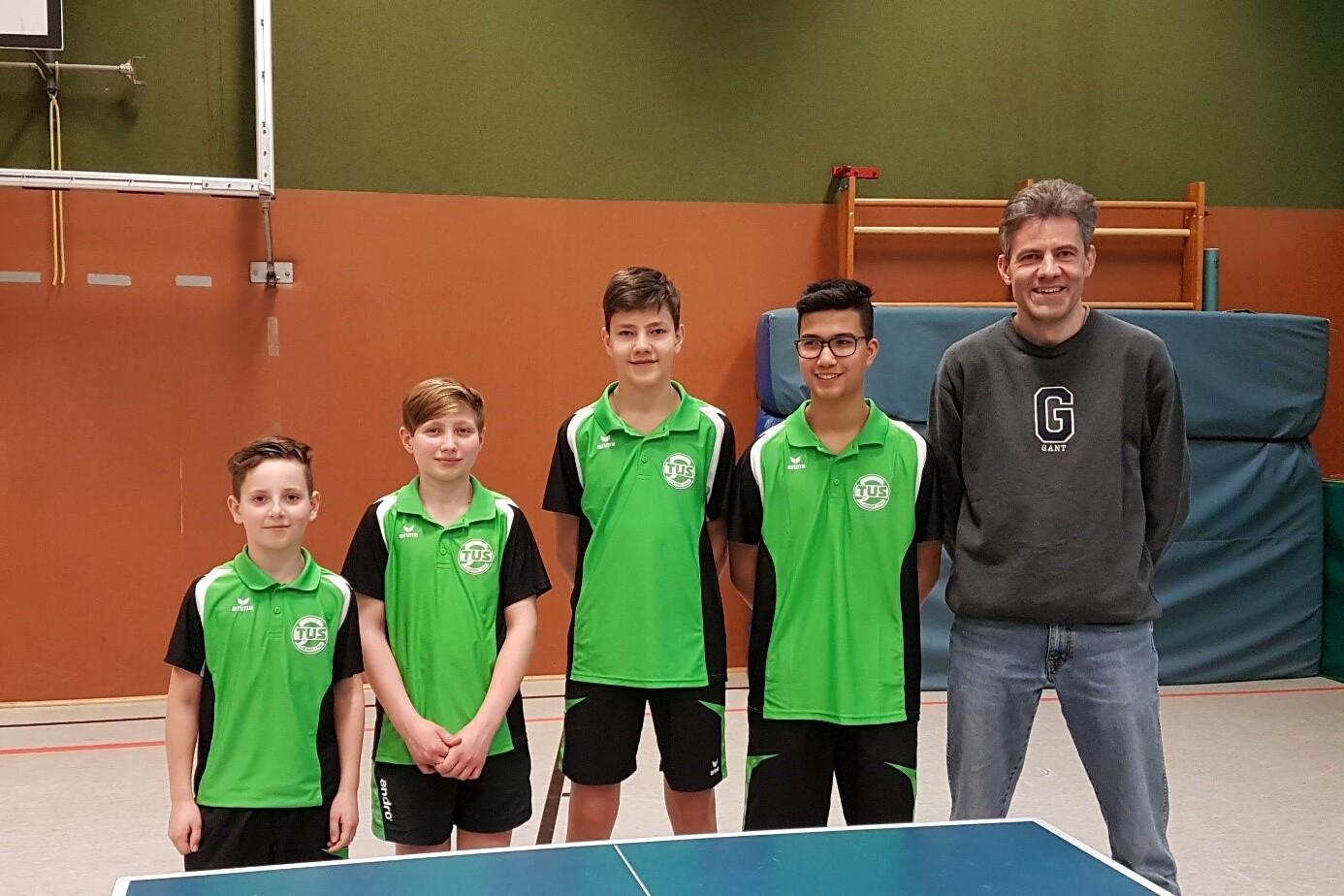 Gruppe von f&uuml;nf Personen in gr&uuml;nen Tischtennis-Trikots stehen neben einem Tischtennistisch in einer Sporthalle.