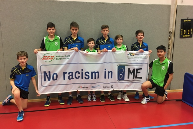 Gruppe von acht Jungen in Sportkleidung h&auml;lt ein Banner mit der Aufschrift "No racism in D ME" in einer Tischtennishalle.