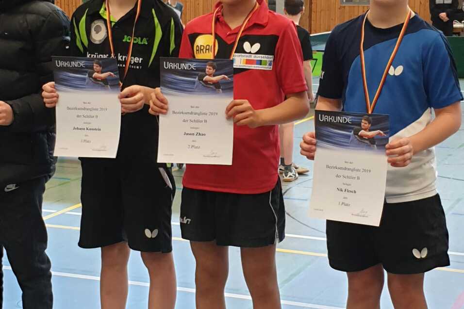 Drei Tischtennisspieler halten ihre Urkunden und Medaillen in der Sporthalle, stehen nebeneinander in Sportbekleidung.