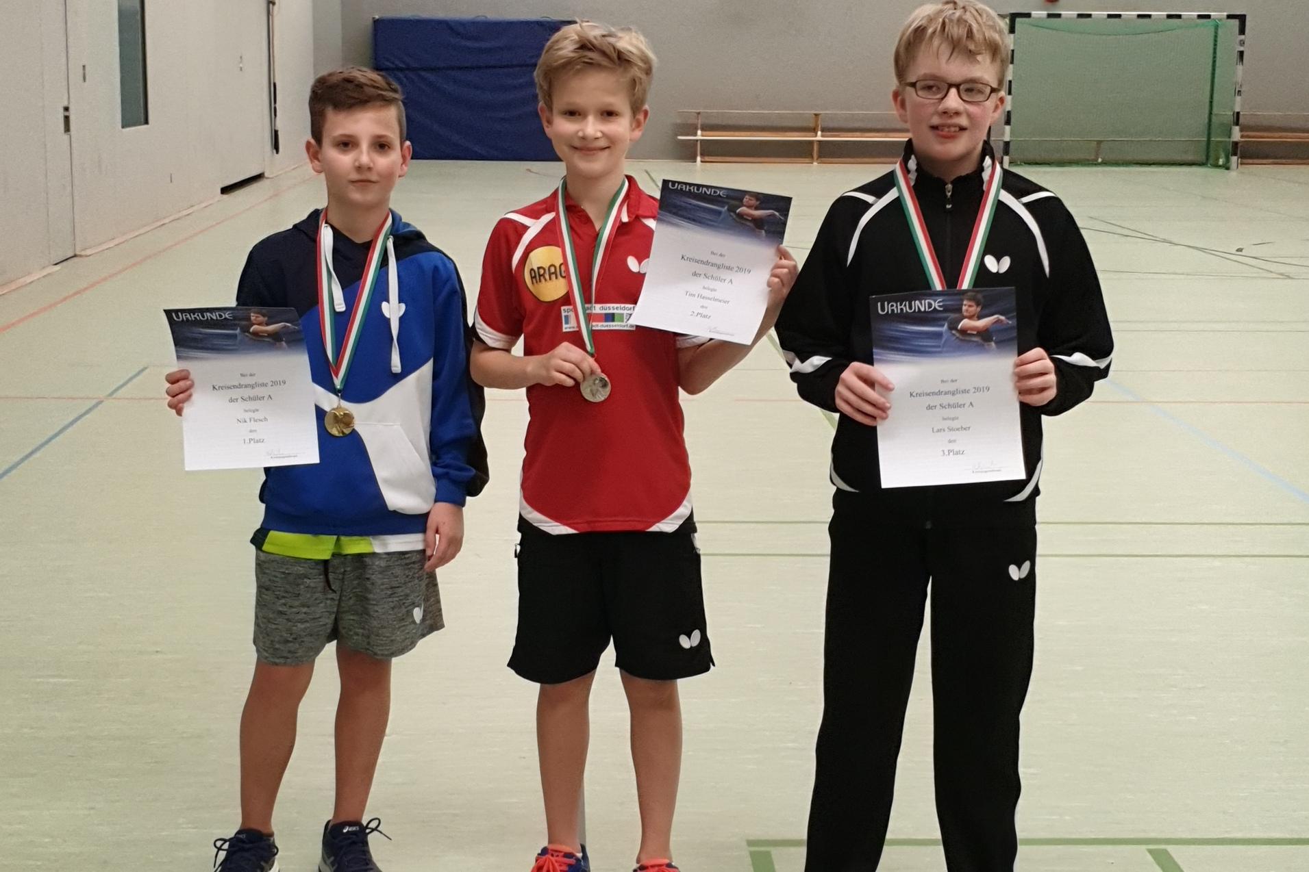 Drei Kinder stehen in einer Sporthalle mit Medaillen und Urkunden in den Händen, lächelnd in Sportkleidung.