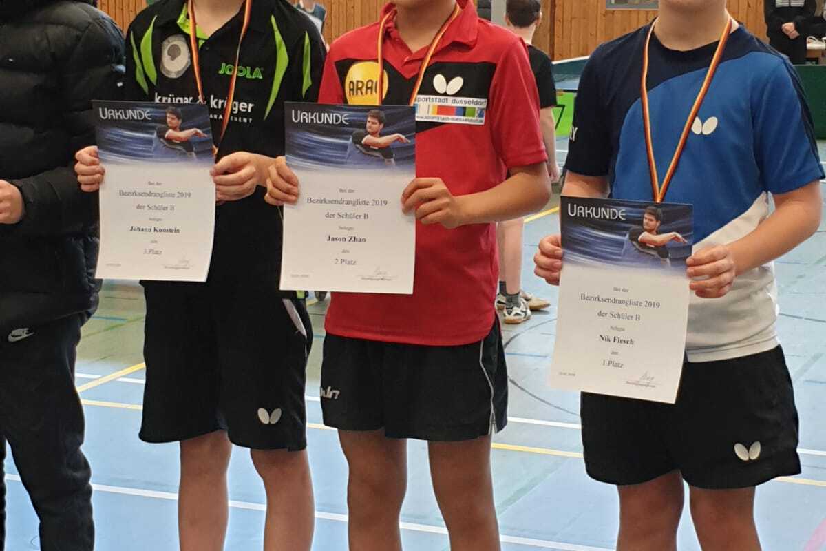 Drei Tischtennisspieler halten ihre Urkunden und Medaillen in der Sporthalle, stehen nebeneinander in Sportbekleidung.