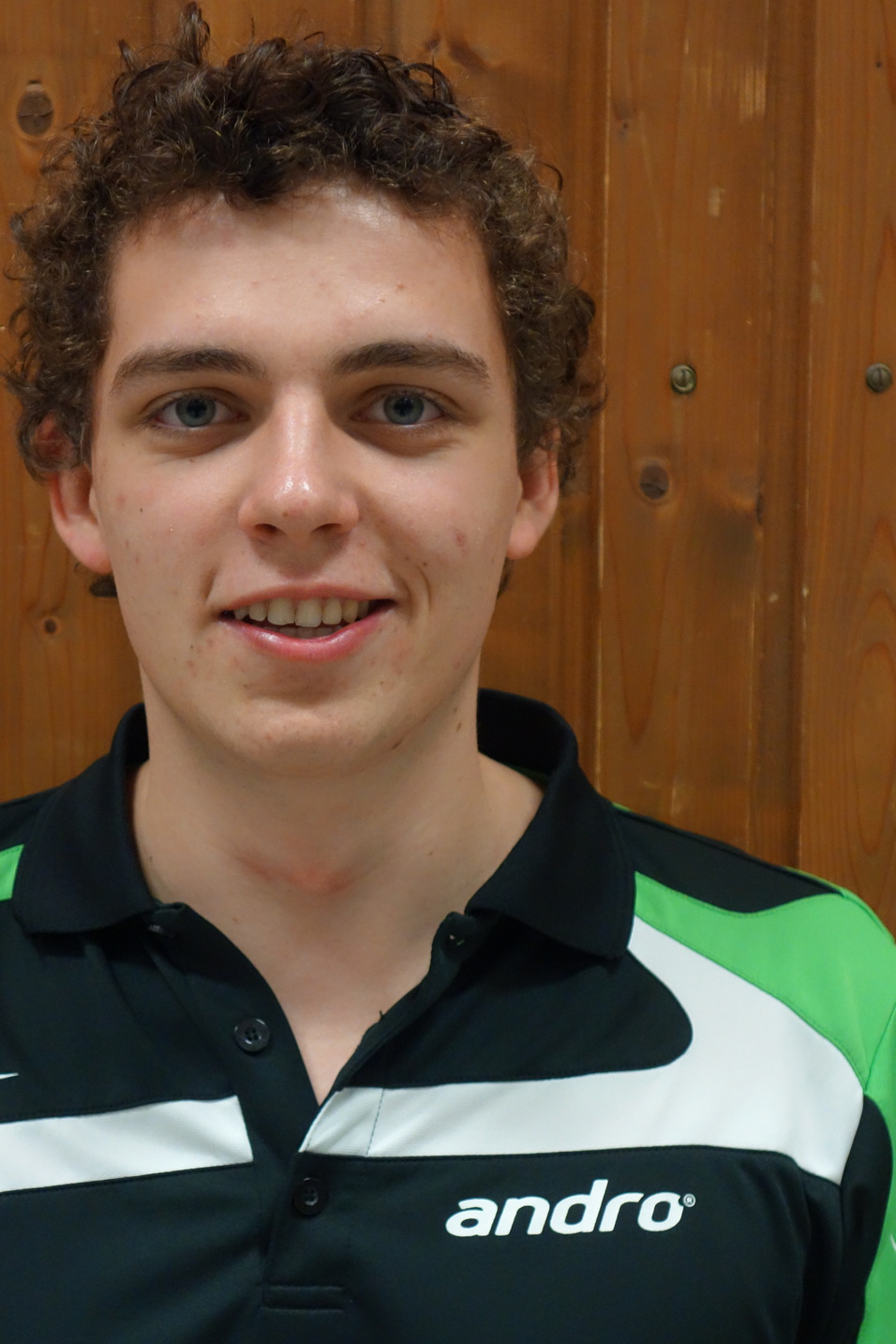 Portr&auml;t eines jungen Mannes mit lockigem Haar, der ein schwarz-gr&uuml;nes Sportshirt tr&auml;gt, vor einer Holzwand.