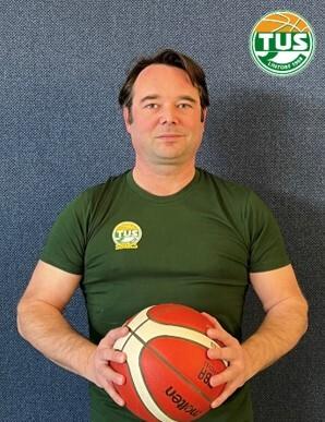 Mann in gr&uuml;nem T-Shirt h&auml;lt einen Basketball vor einem blauen Hintergrund mit Logo oben rechts.