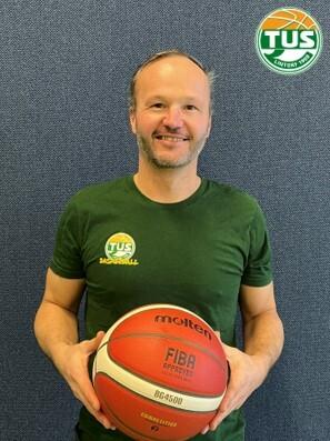 Mann mit Basketball in der Hand, tr&auml;gt ein gr&uuml;nes T-Shirt mit Logo des TuS, vor blauer Wand.