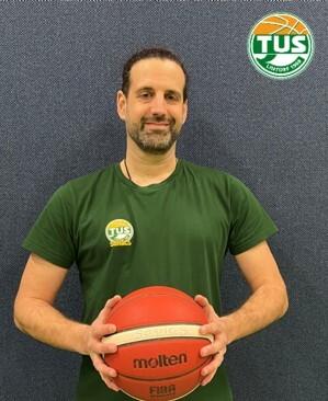 Ein Trainer in einem gr&uuml;nen T-Shirt h&auml;lt einen Basketball mit beiden H&auml;nden vor einem blauen Hintergrund.