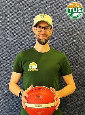 Mann mit Brille und Kap ein T-Shirt mit dem Logo des TUS, h&auml;lt einen Basketball vor einer blauen Wand.