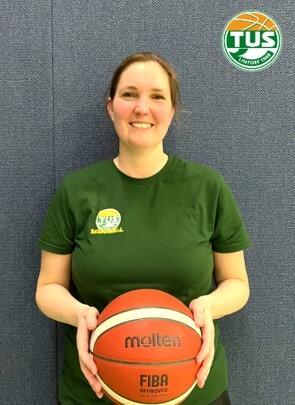 Frau in gr&uuml;nem T-Shirt h&auml;lt einen Basketball vor einer blauen Wand. Logo von TUS oben rechts im Bild sichtbar.