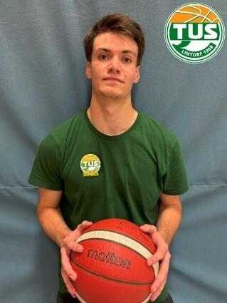 Junger Mann in grünem T-Shirt hält einen Basketball vor einer blauen Wand. Logo des Vereins oben rechts.