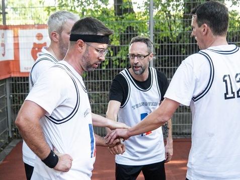 F&uuml;nf Basketballspieler in Trikots bilden einen Zusammenschluss mit H&auml;nden auf einem Sportplatz im Hintergrund.