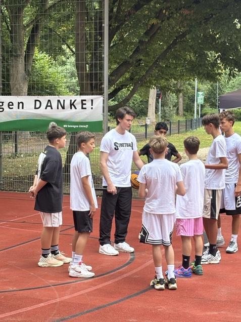 Gruppe von Kindern und Jugendlichen um einen Trainer auf einem Basketballplatz, mit einem Banner im Hintergrund.