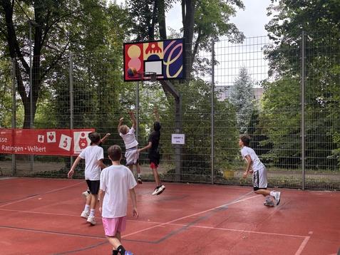 F&uuml;nf Kinder spielen Basketball auf einem Platz, einer wirft den Ball auf den Korb, mehrere B&auml;ume im Hintergrund.