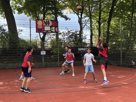 Sechs Kinder spielen Basketball auf einem Sportplatz mit Korbanlage im Hintergrund unter B&auml;umen.