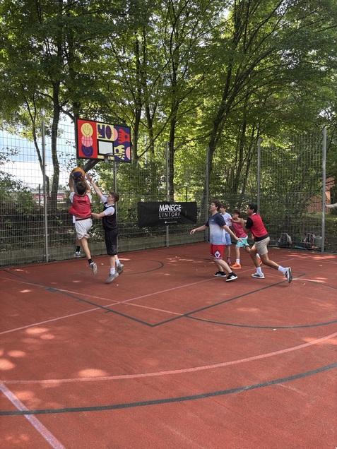 Sechsk&ouml;pfige Gruppe von Kindern spielt Basketball auf einem Au&szlig;ensportplatz, einer springt zum Korb.
