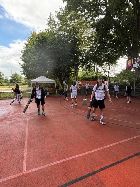 Gruppen von Menschen reinigen einen Basketballplatz mit speziellen Ger&auml;ten unter einem bew&ouml;lkten Himmel.