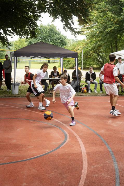 Vier Kinder spielen Basketball auf einem roten Platz, w&auml;hrend Zuschauer hinter einer Markise sitzen.