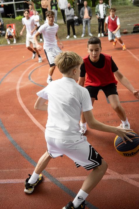 Zwei Jungen spielen Basketball auf einem Sportplatz, Zuschauer stehen im Hintergrund. Ein Junge dribbelt den Ball.