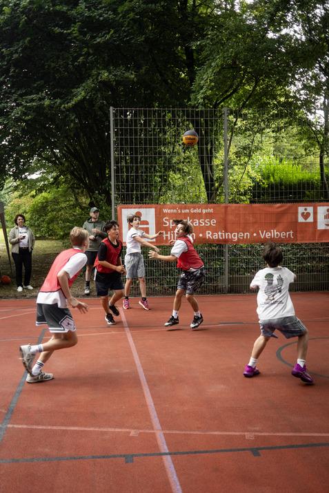 Vier Kinder spielen Basketball auf einem Platz, w&auml;hrend Zuschauer im Hintergrund stehen und zuschauen.
