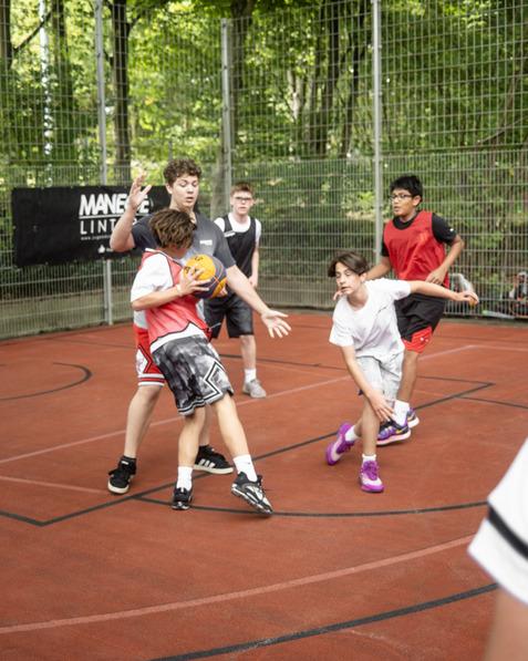 Spielende Kinder auf einem Basketballplatz, zwei Spieler im Kampf um den Ball, umringt von weiteren Mitspielern.