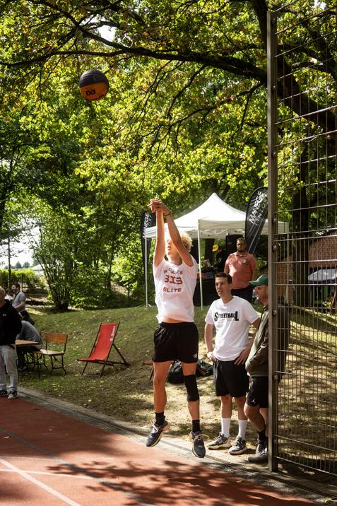 Junger Basketballspieler springt und wirft den Ball Richtung Korb, im Hintergrund Zuschauer und B&auml;ume.