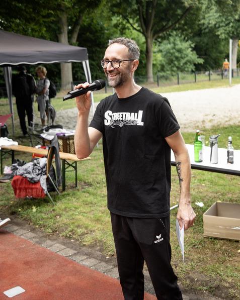 Mann mit Brille und Mikrofon spricht auf einer Veranstaltung im Freien, im Hintergrund sind Tische und Zuschauer zu sehen.