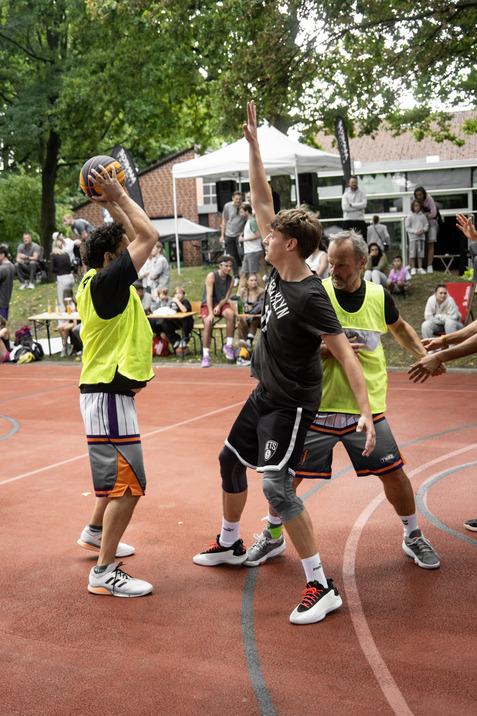 Zwei Basketballspieler in gelben Westen k&auml;mpfen um den Ball auf einem roten Basketballplatz, w&auml;hrend Zuschauer beobachten.