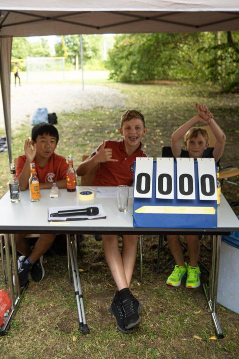 Drei Kinder in roten T-Shirts sitzen an einem Tisch mit einem Punktestand von 0:0 und l&auml;cheln in die Kamera.
