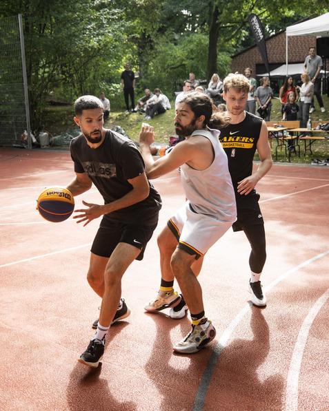 Zwei Basketballspieler k&auml;mpfen um den Ball auf einem Freiplatz, ein Zuschauer beobachtet das Spiel im Hintergrund.