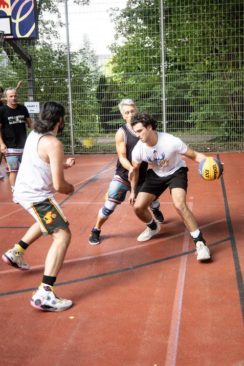 Drei M&auml;nner spielen Basketball auf einem Au&szlig;enspielfeld, w&auml;hrend ein Spieler den Ball dribbelt und verteidigt wird.