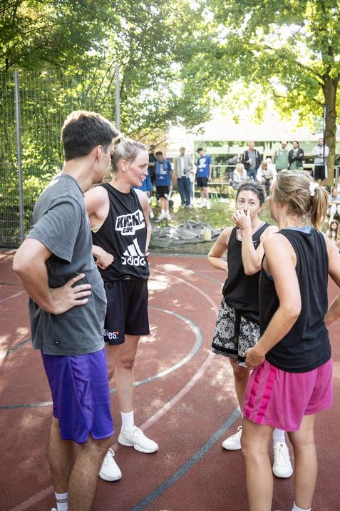 Vier Basketballspieler in sportlicher Kleidung diskutieren auf einem Platz unter B&auml;umen. Zuschauer sind im Hintergrund.