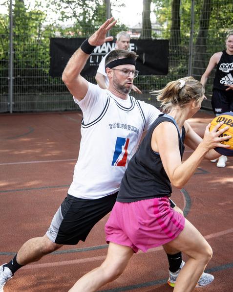 Paar beim Basketballspiel: Ein Mann versucht, den Ball einer Frau abzunehmen, w&auml;hrend andere Spieler im Hintergrund aktiv sind.