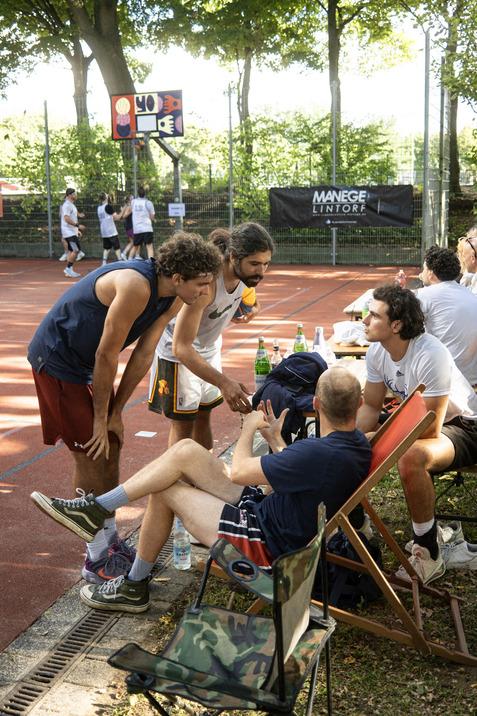 Vier M&auml;nner unterhalten sich auf einem Basketballplatz, w&auml;hrend im Hintergrund Basketballspieler aktiv sind.