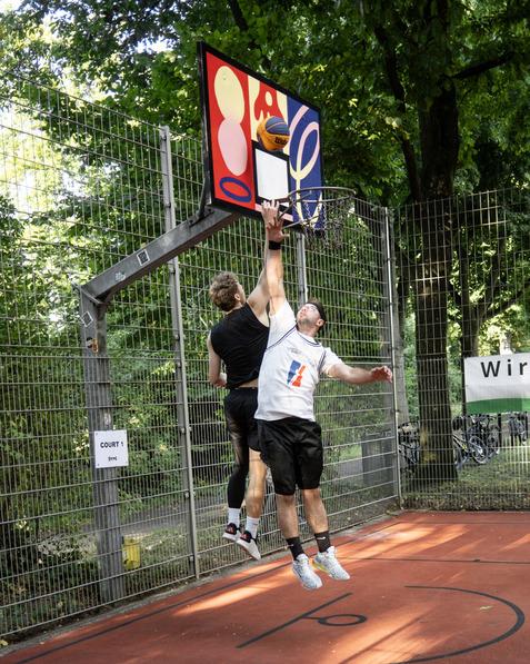 Zwei Basketballspieler springen unter einem Korb, w&auml;hrend einer den Ball fliegt und der andere ihn zu blocken versucht.