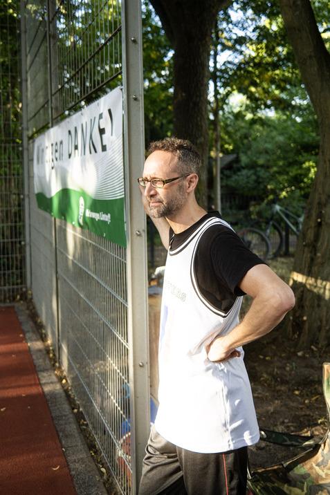 Mann in Sportshirt steht neben einem Zaun mit einem Banner in einem gr&uuml;nen Park. Tageslicht filtert durch die B&auml;ume.