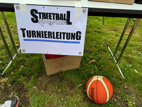 Schild mit der Aufschrift &bdquo;Streetball Turnierleitung&ldquo; und einem Basketball auf dem Boden vor einem Tisch.