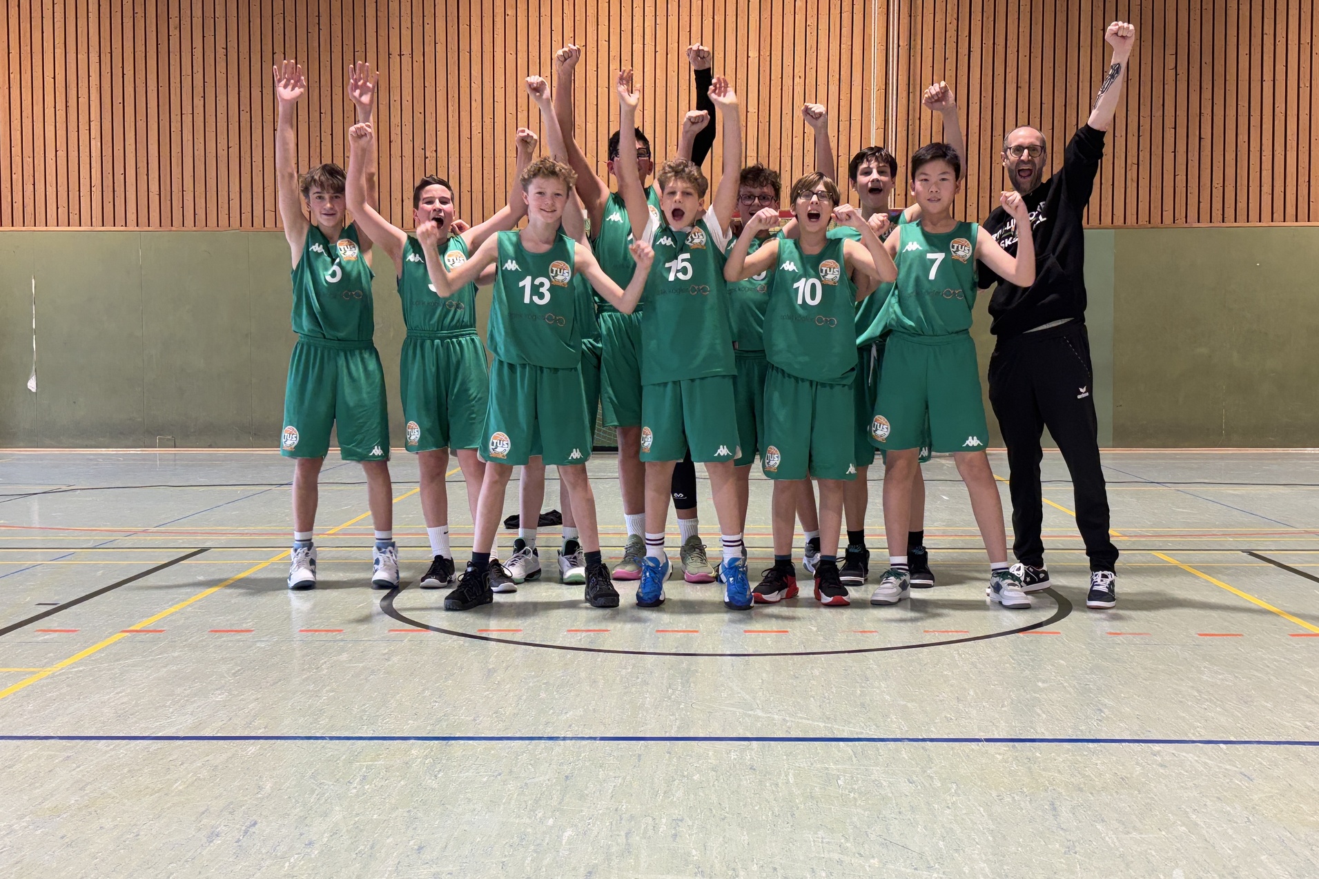 Gruppe jubelnder Basketballspieler in gr&uuml;nen Trikots steht auf einem Sportplatz und hebt die Arme in die Luft.