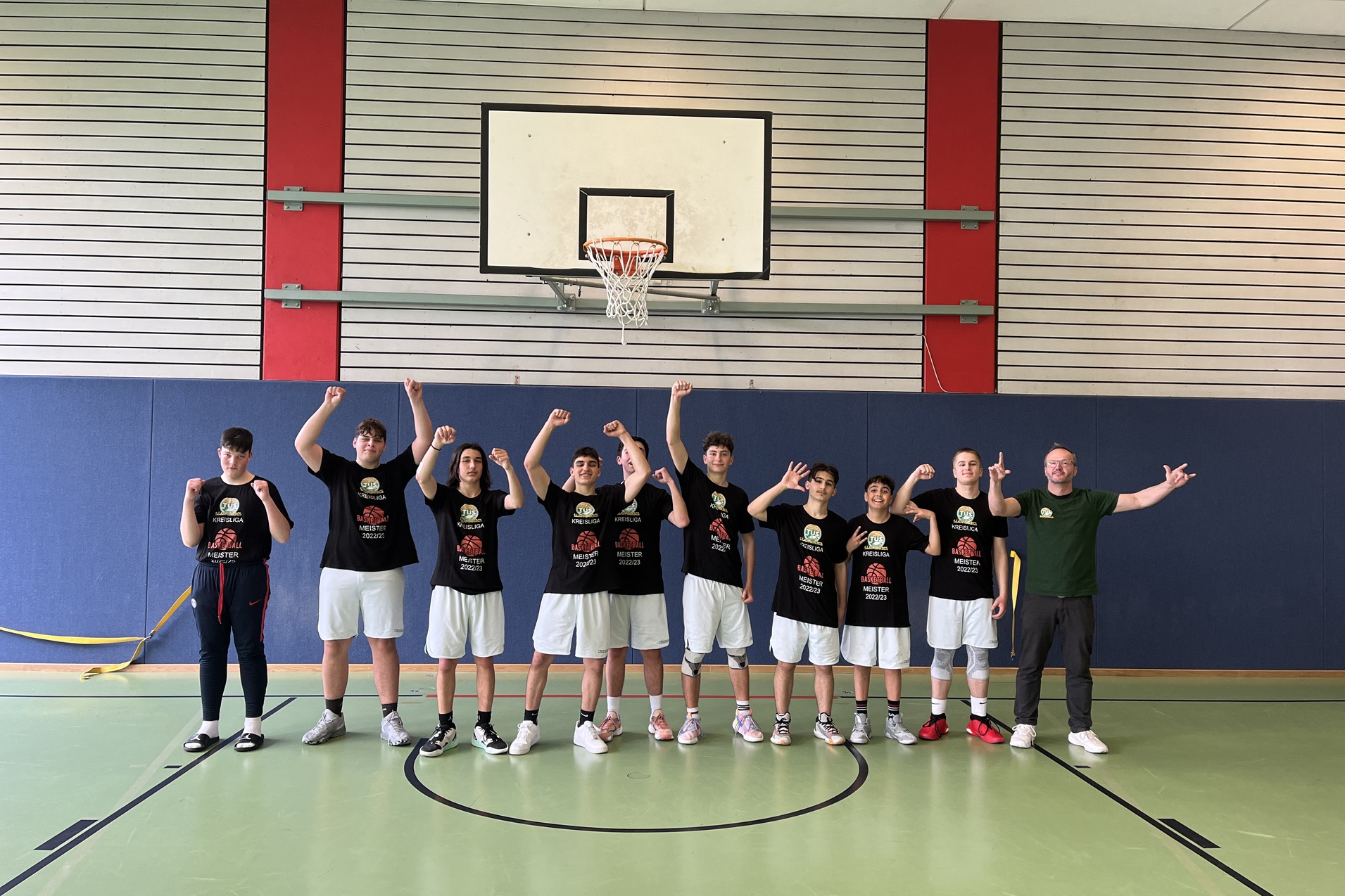 Gruppe von zehn jungen Basketballspielern in einheitlichen schwarzen T-Shirts posiert fr&ouml;hlich auf dem Spielfeld.