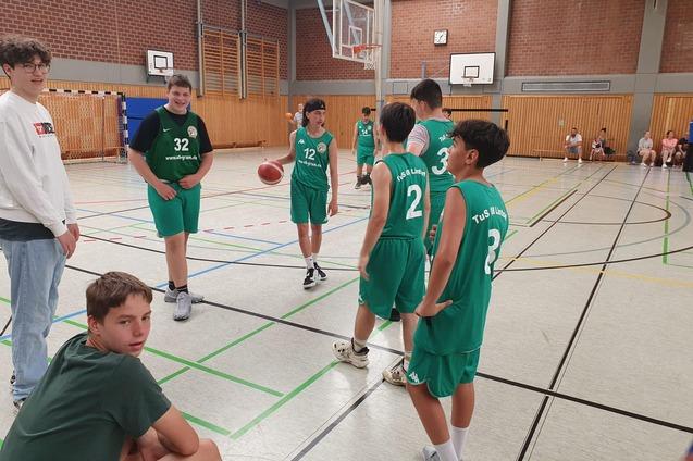 Sechs Jugendliche in gr&uuml;nen Basketballtrikots stehen in einer Sporthalle, einer h&auml;lt den Ball in der Hand.